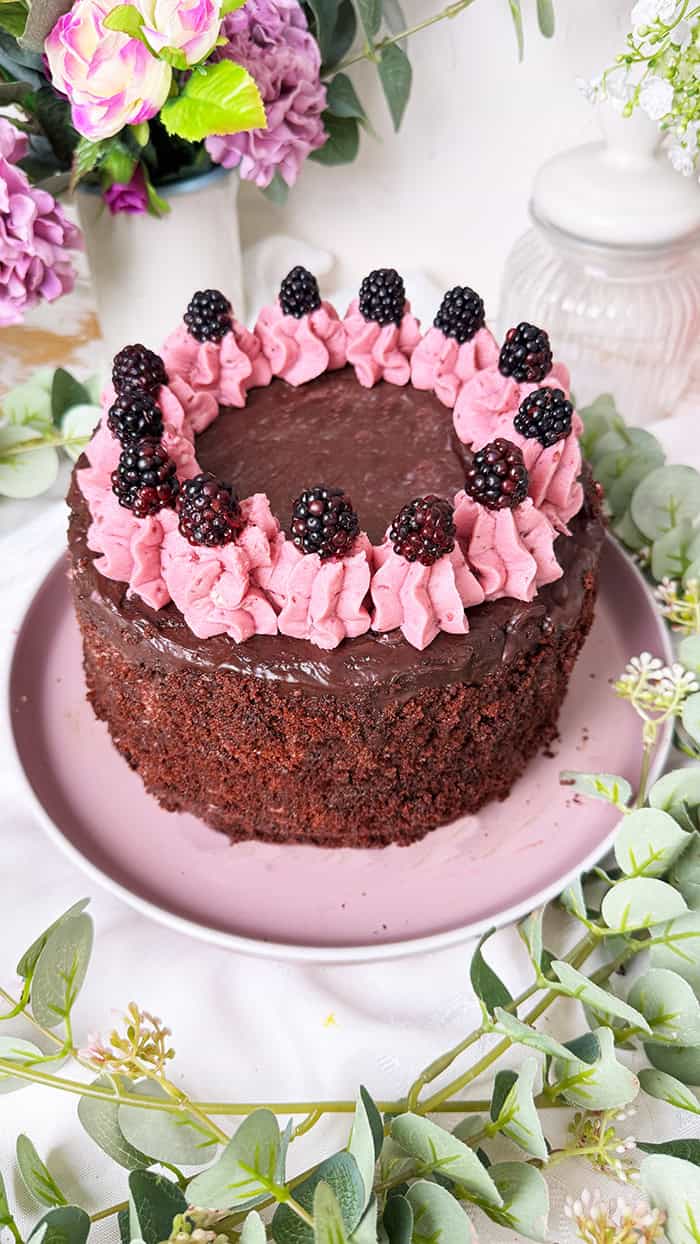 Fruchtige Brombeer-Schokoladen-Torte mit saftigem Schokoboden, pinker Brombeercreme und Ganache-Topping, dekoriert mit frischen Brombeeren, auf rosa Kuchenteller vor Blumenhintergrund. 