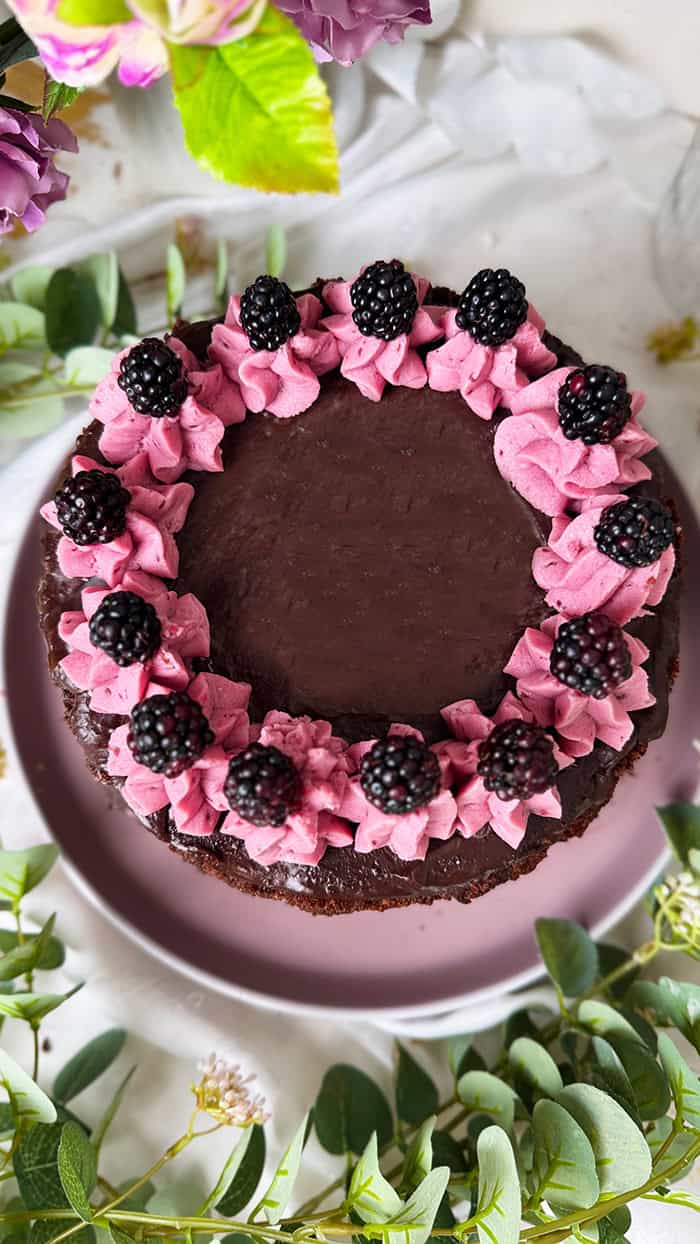 Fruchtige Brombeer-Schokoladen-Torte mit saftigem Schokoboden, pinker Brombeercreme und Ganache-Topping, dekoriert mit frischen Brombeeren, auf rosa Kuchenteller vor Blumenhintergrund. 