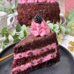 Fruchtige Brombeer-Schokoladen-Torte mit saftigem Schokoboden, pinker Brombeercreme und Ganache-Topping, dekoriert mit frischen Brombeeren, auf rosa Kuchenteller vor Blumenhintergrund.