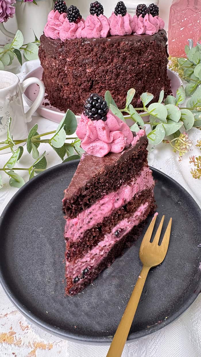 Fruchtige Brombeer-Schokoladen-Torte mit saftigem Schokoboden, pinker Brombeercreme und Ganache-Topping, dekoriert mit frischen Brombeeren, auf rosa Kuchenteller vor Blumenhintergrund. 