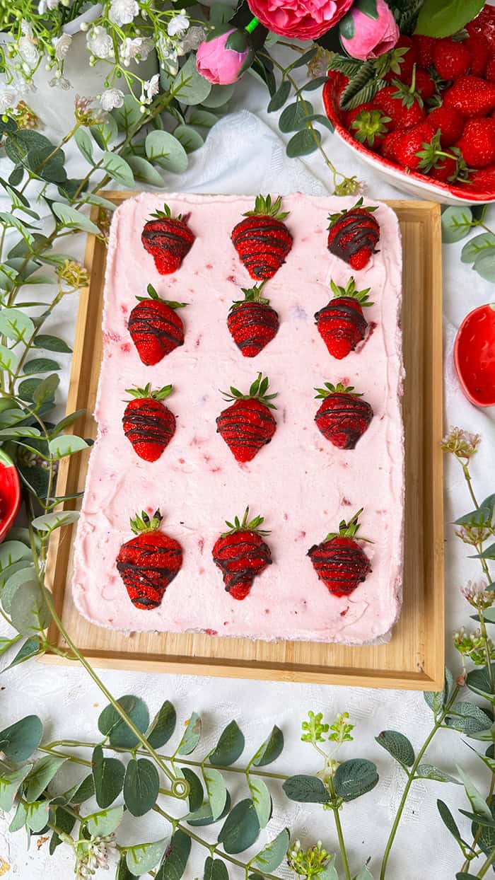 Ein Stück Erdbeeren-Schoko-Kuchen vom Blech mit cremiger Erdbeer-Joghurf-Sahne und frischen Erdbeeren auf einem saftigen Schokobiskuitboden, dekoriert mit Schoko-Erdbeeren und serviert auf einem Teller mit goldener Gabel. 