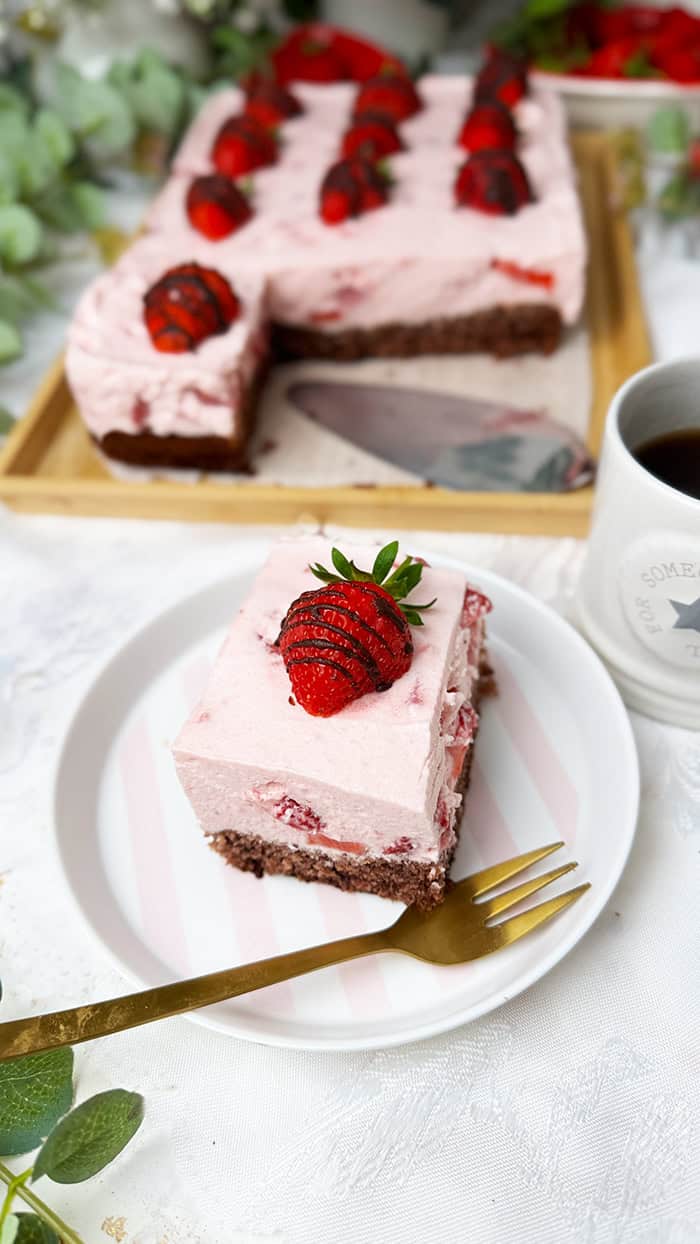 Ein Stück Erdbeeren-Schoko-Kuchen vom Blech mit cremiger Erdbeer-Joghurt-Sahne und frischen Erdbeeren auf einem saftigen Schokobiskuitboden, dekoriert mit Schoko-Erdbeeren und serviert auf einem Teller mit goldener Gabel.