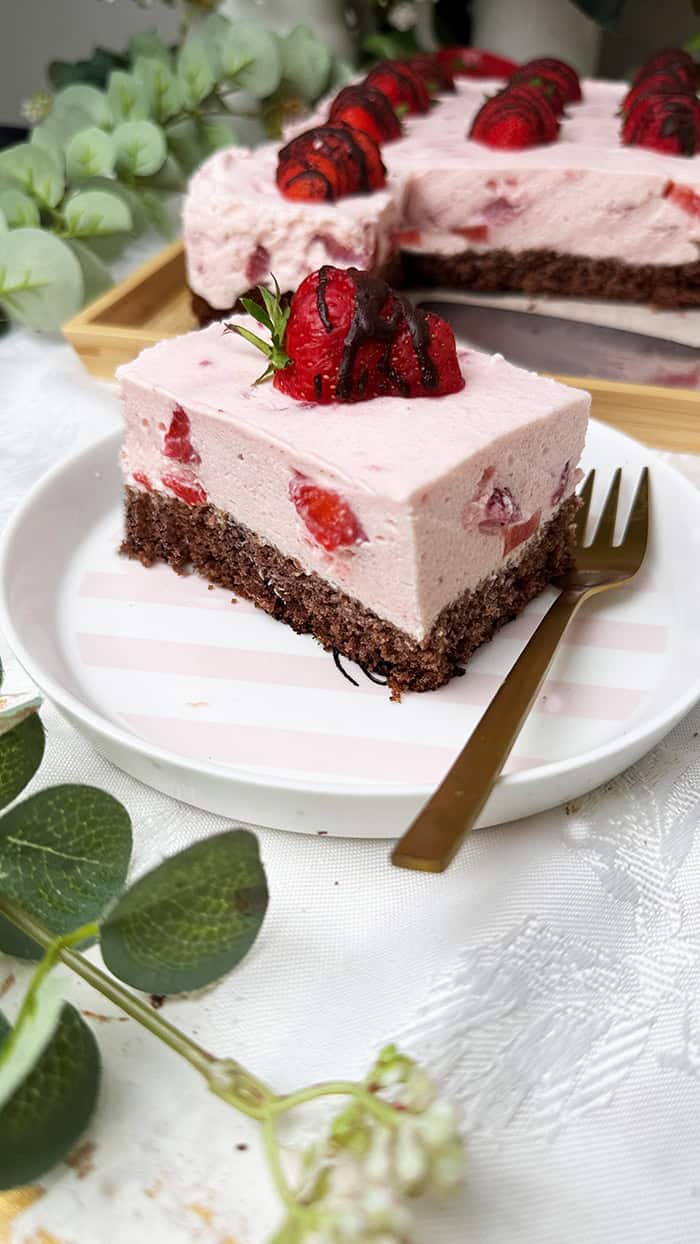 Ein Stück Erdbeeren-Schoko-Kuchen vom Blech mit cremiger Erdbeer-Joghurt-Sahne und frischen Erdbeeren auf einem saftigen Schokobiskuitboden, dekoriert mit Schoko-Erdbeeren und serviert auf einem Teller mit goldener Gabel.