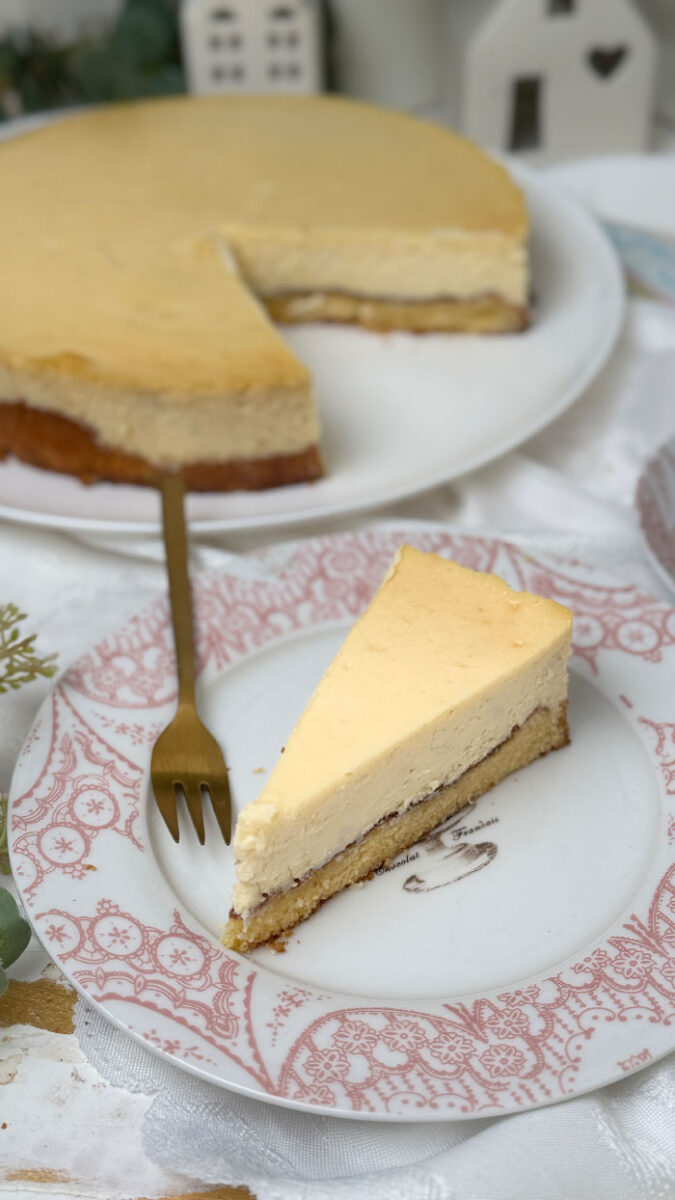 Ein frisch gebackener, goldgelber Marzipan-Cheesecake auf einem weißen Teller. Im Hintergrund eine helle, gemütliche Dekoration mit kleinen weißen Keramikhäusern, grünen Eukalyptuszweigen und weißen Blumen