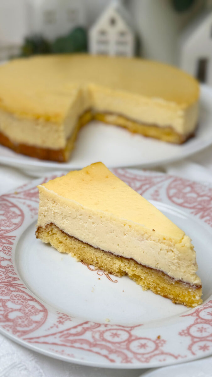 Ein frisch gebackener, goldgelber Marzipan-Cheesecake auf einem weißen Teller. Im Hintergrund eine helle, gemütliche Dekoration mit kleinen weißen Keramikhäusern, grünen Eukalyptuszweigen und weißen Blumen