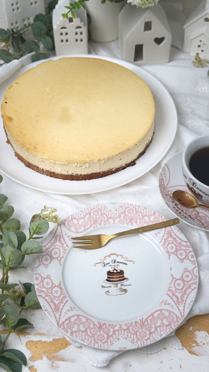 Ein frisch gebackener, goldgelber Marzipan-Cheesecake auf einem weißen Teller. Im Hintergrund eine helle, gemütliche Dekoration mit kleinen weißen Keramikhäusern, grünen Eukalyptuszweigen und weißen Blumen