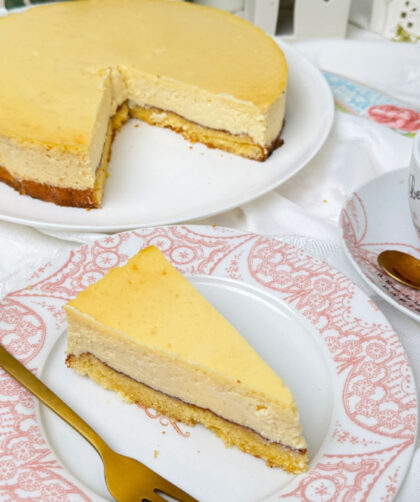 Ein frisch gebackener, goldgelber Marzipan-Cheesecake auf einem weißen Teller. Im Hintergrund eine helle, gemütliche Dekoration mit kleinen weißen Keramikhäusern, grünen Eukalyptuszweigen und weißen Blumen