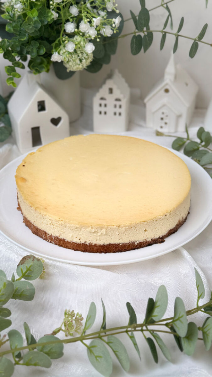 Ein frisch gebackener, goldgelber Marzipan-Cheesecake auf einem weißen Teller. Im Hintergrund eine helle, gemütliche Dekoration mit kleinen weißen Keramikhäusern, grünen Eukalyptuszweigen und weißen Blumen