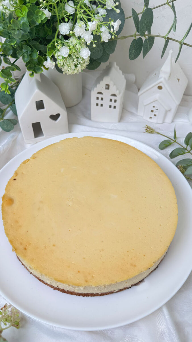 Ein frisch gebackener, goldgelber Marzipan-Cheesecake auf einem weißen Teller. Im Hintergrund eine helle, gemütliche Dekoration mit kleinen weißen Keramikhäusern, grünen Eukalyptuszweigen und weißen Blumen