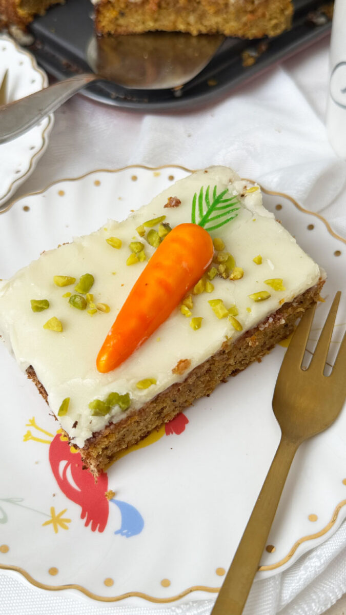 Schweizer Rüblikuchen: Das Original-Rezept für den einfachen saftigen Klassiker 6 Saftiges Stück Schweizer Rüblikuchen mit weißem Zuckerguss, Pistazien und einer Marzipanrübe auf einem weißen Teller mit Goldrand