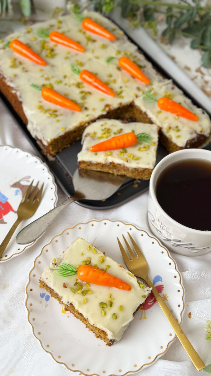 Schweizer Rüblikuchen: Das Original-Rezept für den einfachen saftigen Klassiker 5 Saftiges Stück Schweizer Rüblikuchen mit weißem Zuckerguss, Pistazien und einer Marzipanrübe auf einem weißen Teller mit Goldrand