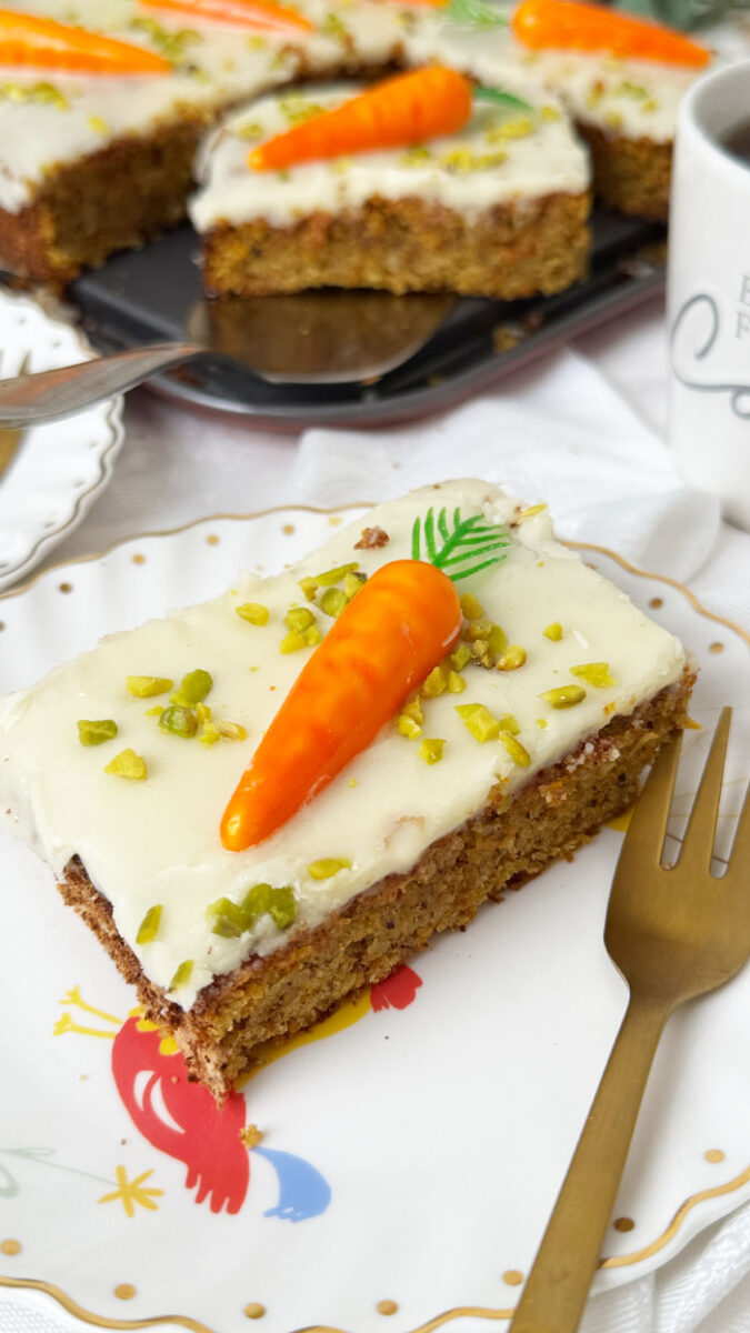 Schweizer Rüblikuchen: Das Original-Rezept für den einfachen saftigen Klassiker 1 Saftiges Stück Schweizer Rüblikuchen mit weißem Zuckerguss, Pistazien und einer Marzipanrübe auf einem weißen Teller mit Goldrand