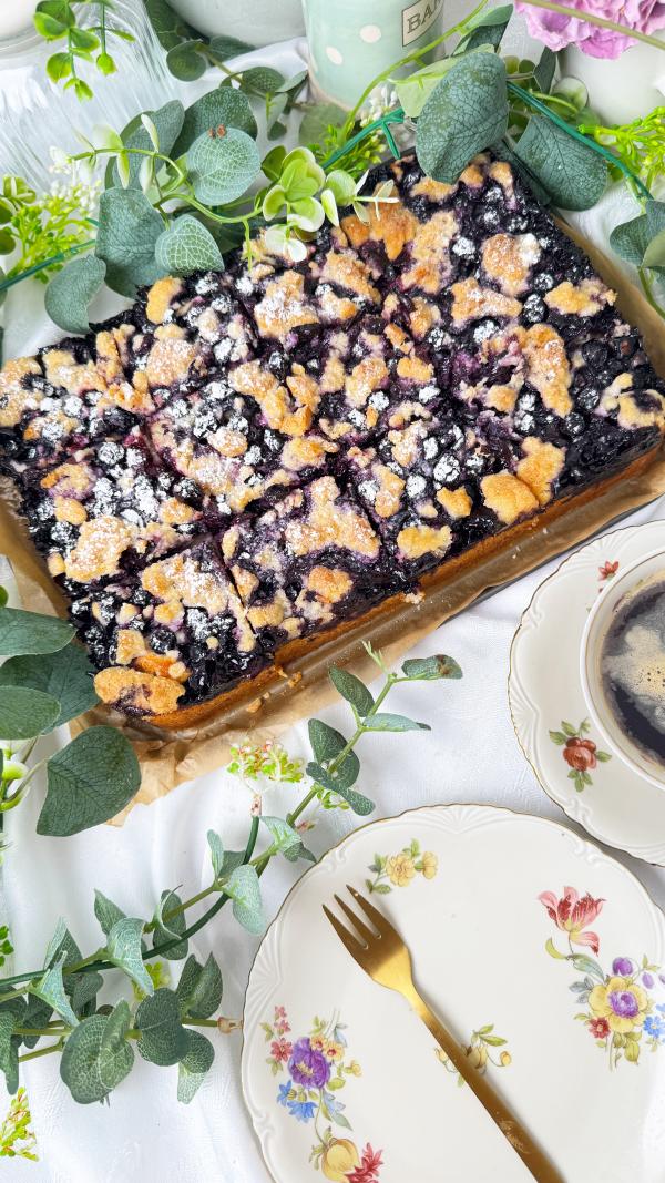 Saftiger Heidelbeerkuchen vom Blech mit Streuseln, Omas Rezept mit Quark-Öl-Teig 6 Ein frisch gebackener Heidelbeerkuchen vom Blech mit Streuseln , umrahmt von Eukalyptuszweigen. Im Vordergrund liegen ein dekorierter Vintage-Teller mit einer goldenen Kuchengabel und eine Tasse Kaffee.