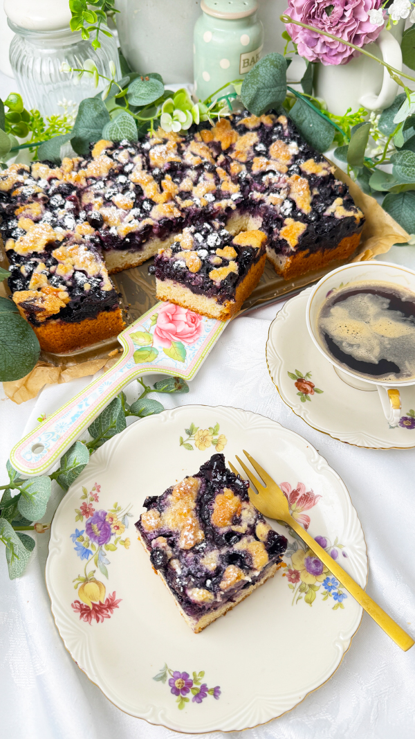 Saftiger Heidelbeerkuchen vom Blech mit Streuseln, Omas Rezept mit Quark-Öl-Teig 1 Ein frisch gebackener, saftiger Heidelbeerkuchen vom Blech mit Streuseln , dekoriert mit Puderzucker. Ein einzelnes Kuchenstück liegt auf einem nostalgischen Blümchenteller mit einer goldenen Kuchengabel daneben, begleitet von einer Tasse Kaffee in einer passenden Vintage-Tasse. Das Setting wird von Eukalyptuszweigen und pastellfarbenen Küchenaccessoires umrahmt.