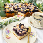 Ein frisch gebackener, saftiger Heidelbeerkuchen vom Blech mit Streuseln , dekoriert mit Puderzucker. Ein einzelnes Kuchenstück liegt auf einem nostalgischen Blümchenteller mit einer goldenen Kuchengabel daneben, begleitet von einer Tasse Kaffee in einer passenden Vintage-Tasse. Das Setting wird von Eukalyptuszweigen und pastellfarbenen Küchenaccessoires umrahmt.