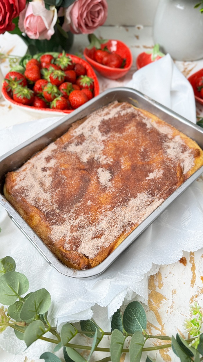 Churros Cheesecake-Schnitte auf einem schwarzen Teller mit einer goldenen Gabel. Die Schnitte hat eine goldbraune, knusprige Zimt-Zucker-Kruste und eine cremige Käsekuchenfüllung. Im Hintergrund steht eine Backform mit dem restlichen Kuchen sowie eine Schüssel mit frischen Erdbeeren und dekorativen Blumen
