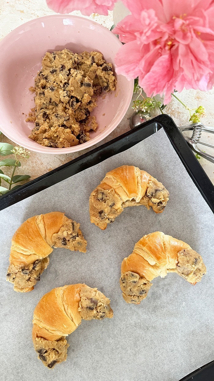 Das Bild zeigt vier Crookies auf einem schwarzen Teller, umgeben von rosa Blumen und Eukalyptuszweigen. Ein Crookie ist eine Kombination aus einem Croissant und einem Cookie. Die Crookies sind halb Croissant mit ihrem goldbraunen, glänzenden Teig und halb Cookie mit ihrem dickeren, bräunlichen Keksanteil, der Schokoladenstücke enthält.