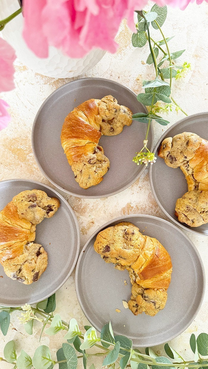 Das Bild zeigt vier Crookies auf einem schwarzen Teller, umgeben von rosa Blumen und Eukalyptuszweigen. Ein Crookie ist eine Kombination aus einem Croissant und einem Cookie. Die Crookies sind halb Croissant mit ihrem goldbraunen, glänzenden Teig und halb Cookie mit ihrem dickeren, bräunlichen Keksanteil, der Schokoladenstücke enthält.
