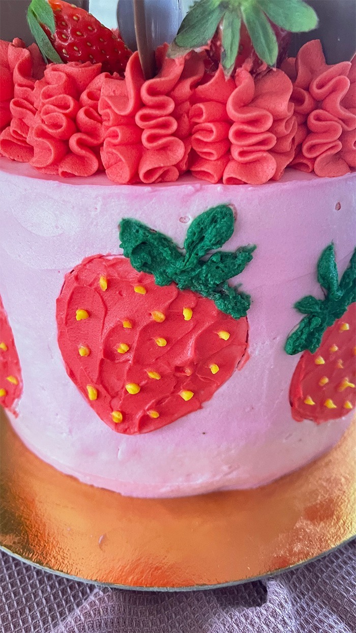 Dies ist ein Bild von einer dekorierten Erdbeer-Schoko-Torte. Die Torte hat eine Basis aus rosa Creme und zeigt mehrere handgefertigte Erdbeerdesigns an den Seiten. Der obere Teil der Torte ist mit rotem Rüschen-Creme, frischen Erdbeeren und Schokoladendekorationen in Herzform verziert. Die Torte steht auf einem goldfarbenen Tortenbrett.