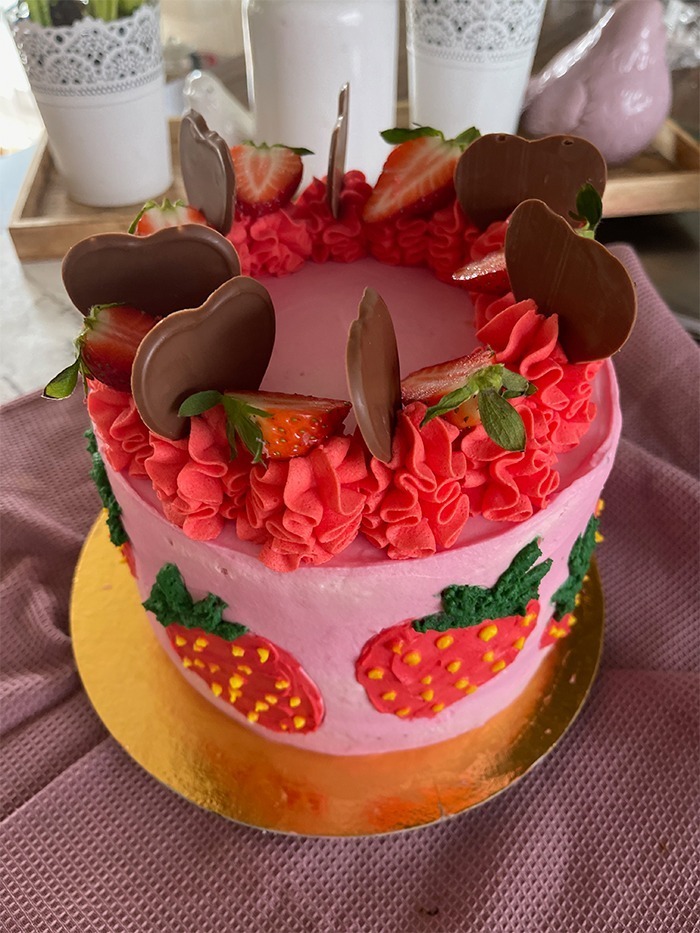 Dies ist ein Bild von einer dekorierten Erdbeer-Schoko-Torte. Die Torte hat eine Basis aus rosa Creme und zeigt mehrere handgefertigte Erdbeerdesigns an den Seiten. Der obere Teil der Torte ist mit rotem Rüschen-Creme, frischen Erdbeeren und Schokoladendekorationen in Herzform verziert. Die Torte steht auf einem goldfarbenen Tortenbrett.