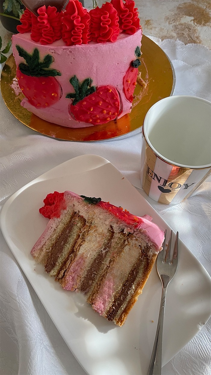 Dies ist ein Bild von einer dekorierten Erdbeer-Schoko-Torte. Die Torte hat eine Basis aus rosa Creme und zeigt mehrere handgefertigte Erdbeerdesigns an den Seiten. Der obere Teil der Torte ist mit rotem Rüschen-Creme, frischen Erdbeeren und Schokoladendekorationen in Herzform verziert. Die Torte steht auf einem goldfarbenen Tortenbrett.
