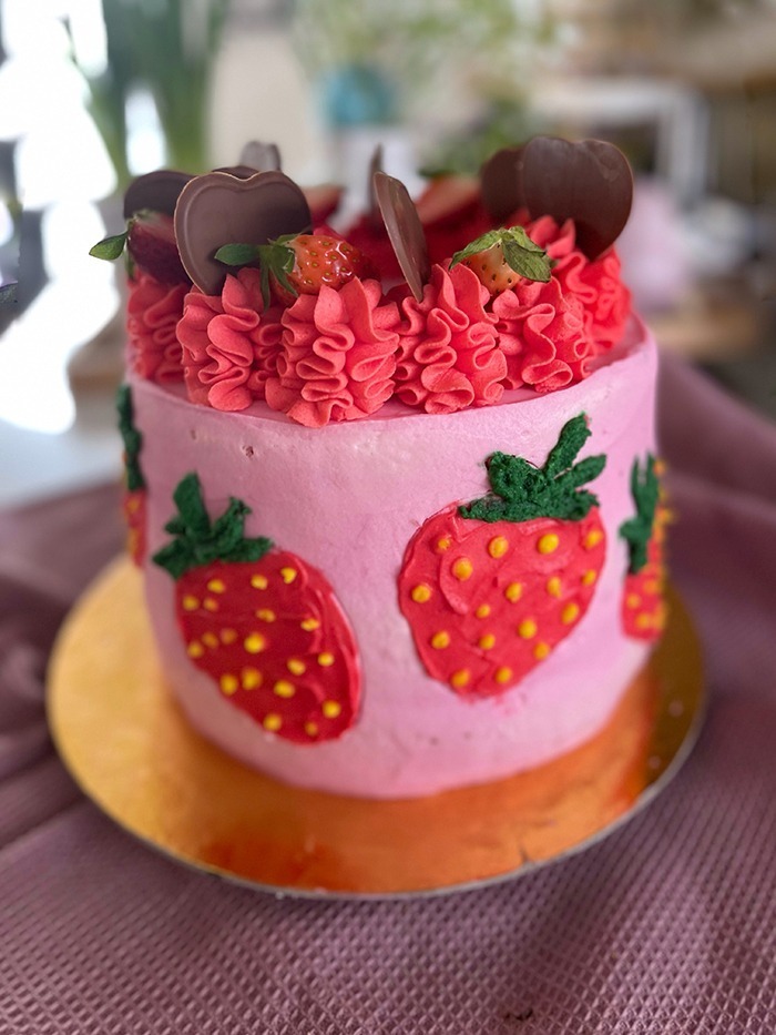 Dies ist ein Bild von einer dekorierten Erdbeer-Schoko-Torte. Die Torte hat eine Basis aus rosa Creme und zeigt mehrere handgefertigte Erdbeerdesigns an den Seiten. Der obere Teil der Torte ist mit rotem Rüschen-Creme, frischen Erdbeeren und Schokoladendekorationen in Herzform verziert. Die Torte steht auf einem goldfarbenen Tortenbrett.
