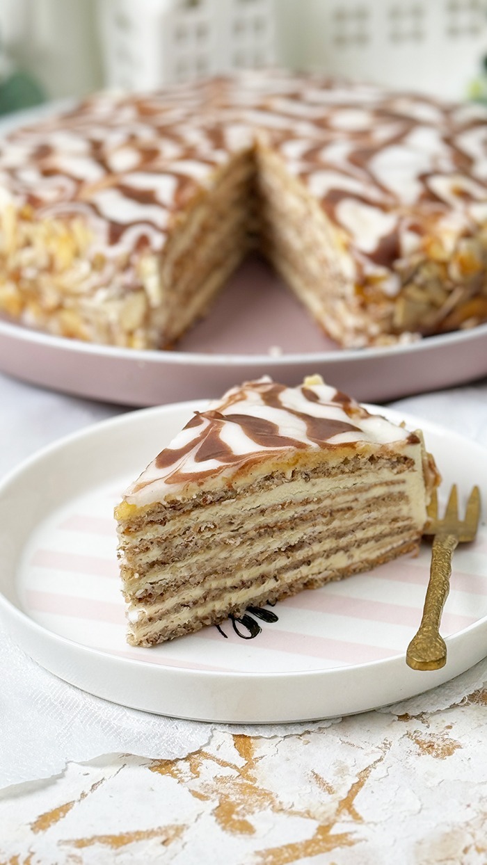 Das Bild zeigt eine Esterházy-Torte, die aus mehreren dünnen Schichten besteht, gefüllt mit Buttercreme, und oben mit einer weißen Glasur und dem typischen Kakaomuster verziert ist.