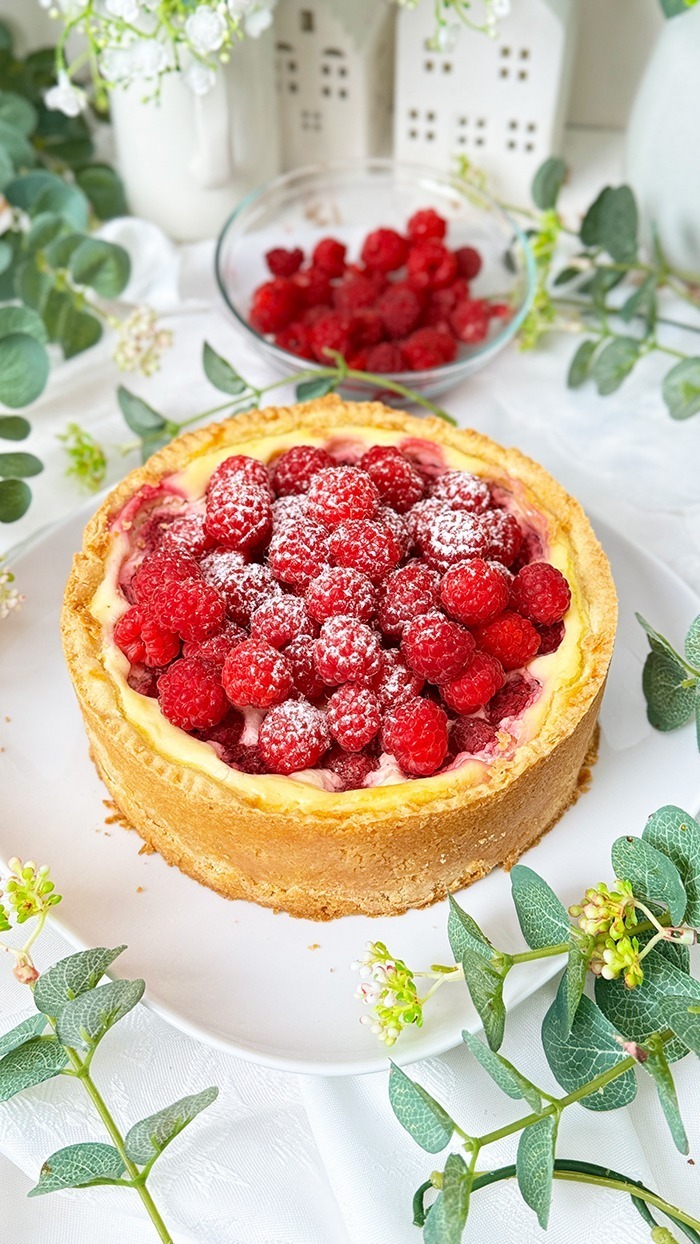 Ein Himbeer-Schmand-Kuchen auf einen Teller garniert mit frischen Himbeeren und leicht mit Puderzucker bestäubt.