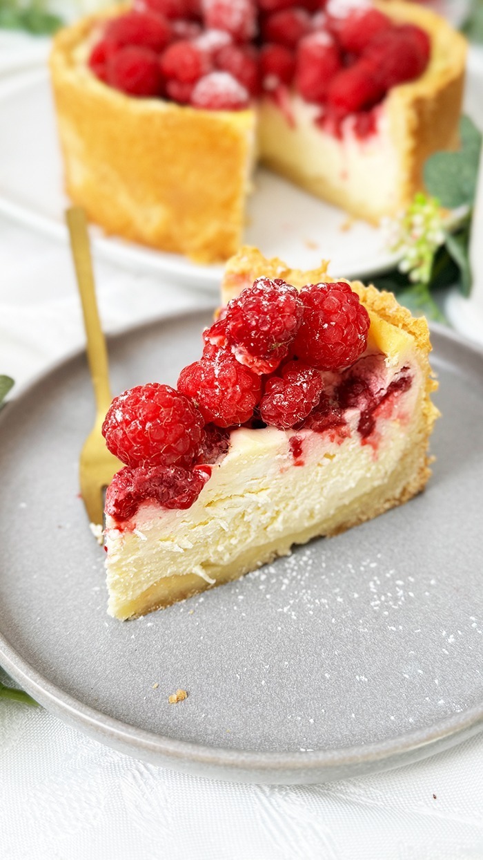 ein Stück Himbeer-Schmand-Kuchen auf einem Teller, garniert mit frischen Himbeeren und leicht mit Puderzucker bestäubt.