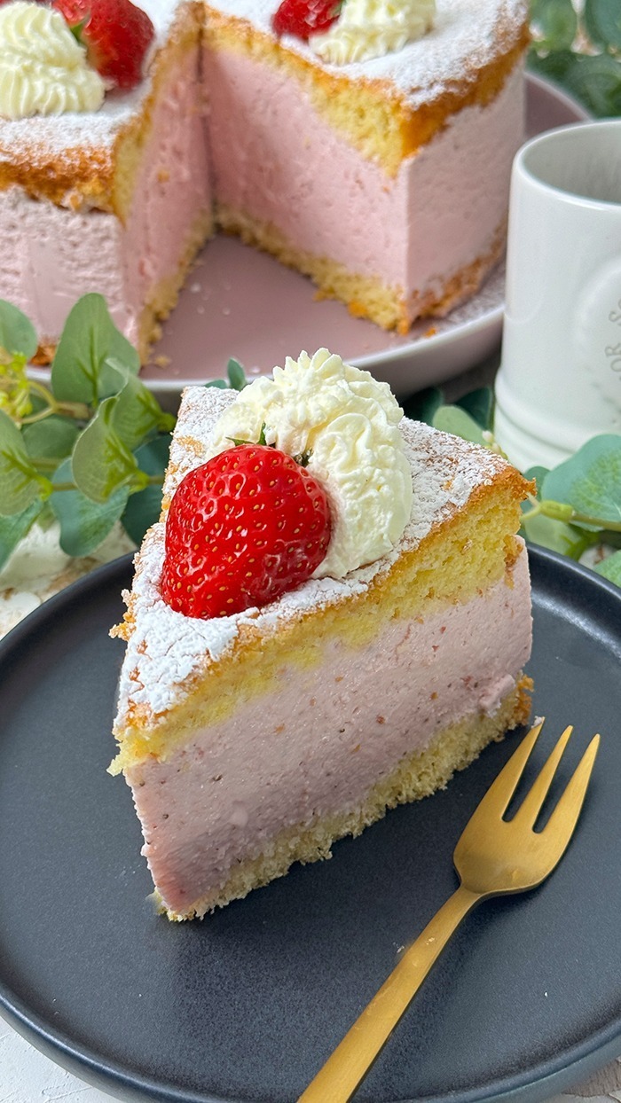 Die Käse-Sahne-Torte mit Erdbeeren hat einen luftigen, goldbraunen Biskuitboden und eine dicke, rosa Käse-Sahne-Füllung mit Erdbeergeschmack. Auf der obersten Schicht sind frische Erdbeeren und Sahnetupfer kunstvoll arrangiert, die mit einer feinen Schicht Puderzucker bestreut sind. Ein bereits geschnittenes Stück zeigt die cremige Füllung und die luftigen Schichten, die die Torte besonders einladend machen.