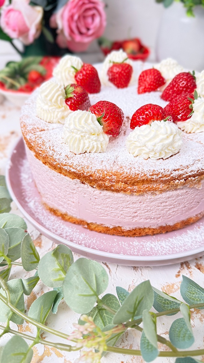 Die Käse-Sahne-Torte mit Erdbeeren hat einen luftigen, goldbraunen Biskuitboden und eine dicke, rosa Käse-Sahne-Füllung mit Erdbeergeschmack. Auf der obersten Schicht sind frische Erdbeeren und Sahnetupfer kunstvoll arrangiert, die mit einer feinen Schicht Puderzucker bestreut sind. Ein bereits geschnittenes Stück zeigt die cremige Füllung und die luftigen Schichten, die die Torte besonders einladend machen.