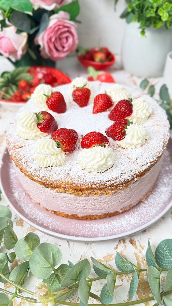 Die Käse-Sahne-Torte mit Erdbeeren hat einen luftigen, goldbraunen Biskuitboden und eine dicke, rosa Käse-Sahne-Füllung mit Erdbeergeschmack. Auf der obersten Schicht sind frische Erdbeeren und Sahnetupfer kunstvoll arrangiert, die mit einer feinen Schicht Puderzucker bestreut sind. Ein bereits geschnittenes Stück zeigt die cremige Füllung und die luftigen Schichten, die die Torte besonders einladend machen.