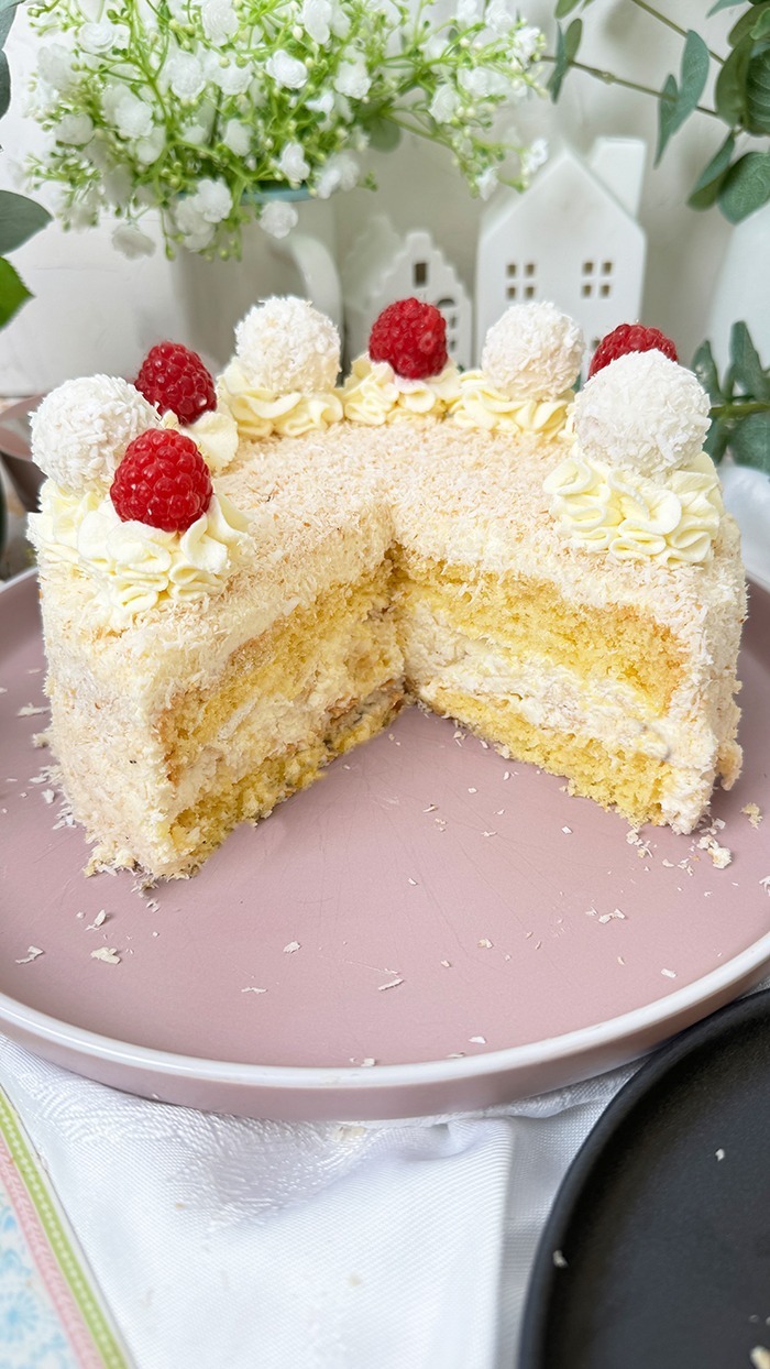 Das Bild zeigt eine Raffaello-Torte mit mehreren Schichten hellem Biskuit und cremiger Füllung, bedeckt mit Kokosraspeln. Die Torte ist dekoriert mit Raffaello-Kugeln, Himbeeren und Sahnetupfen. Ein Stück Torte liegt auf einem grauen Teller mit einer goldenen Gabel daneben, umgeben von grünen Pflanzen und weißen Blumen.