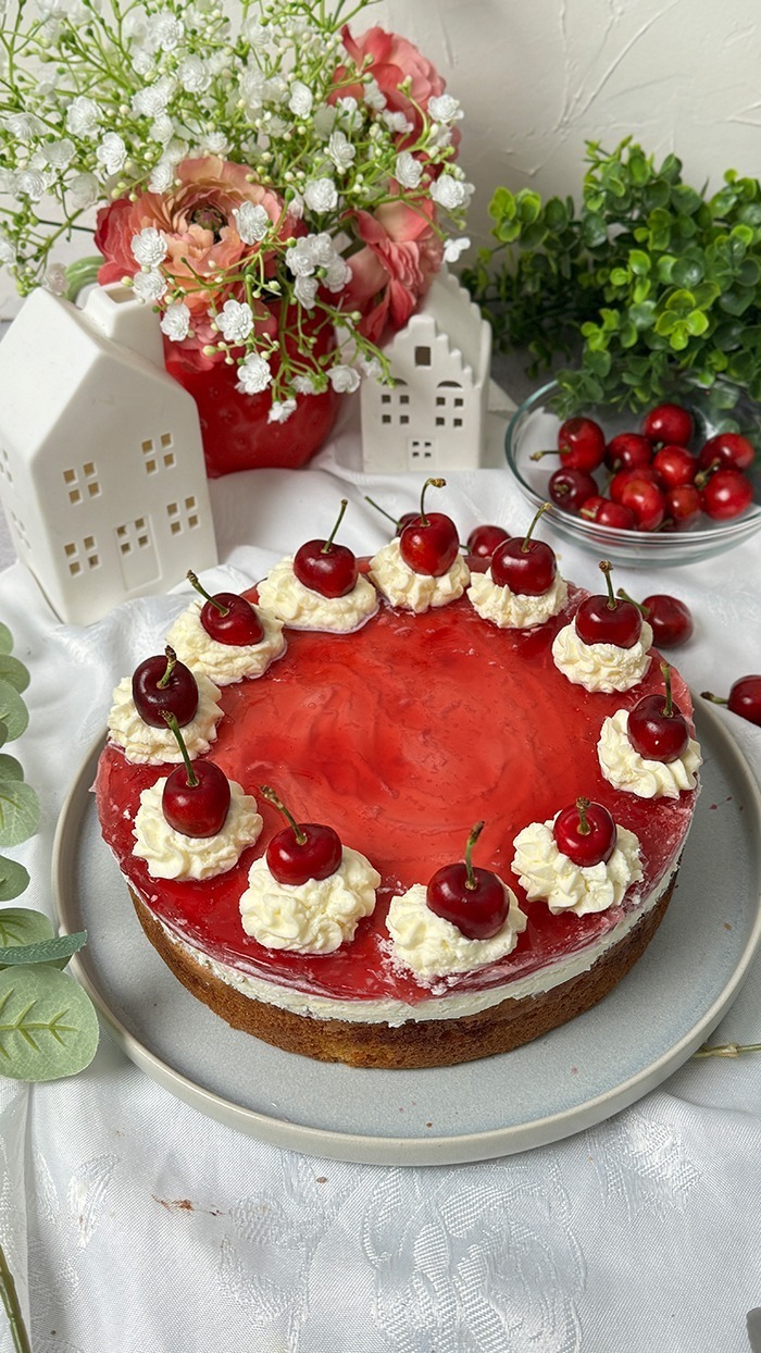 Auf dem Bild sieht man einen Rotkäppchen Kuchen, der auf einem Teller präsentiert wird. Der Kuchen hat eine helle, leicht goldene Biskuitbasis, die mit einer Schicht Sahne oder Pudding und einer roten Geleeschicht bedeckt ist. Am Rand des Kuchens befinden sich Sahnetupfen, auf denen jeweils eine frische, glänzende Kirsche mit Stiel platziert ist. Die Sahnetupfen sind gleichmäßig rund um den Kuchen verteilt. Im Hintergrund sieht man eine stilvolle Dekoration mit kleinen weißen Häuschen und einem Blumenstrauß mit weißen und rosafarbenen Blüten. Die Szene ist auf einem weißen Tischtuch arrangiert, was dem Ganzen eine elegante und einladende Atmosphäre verleiht.