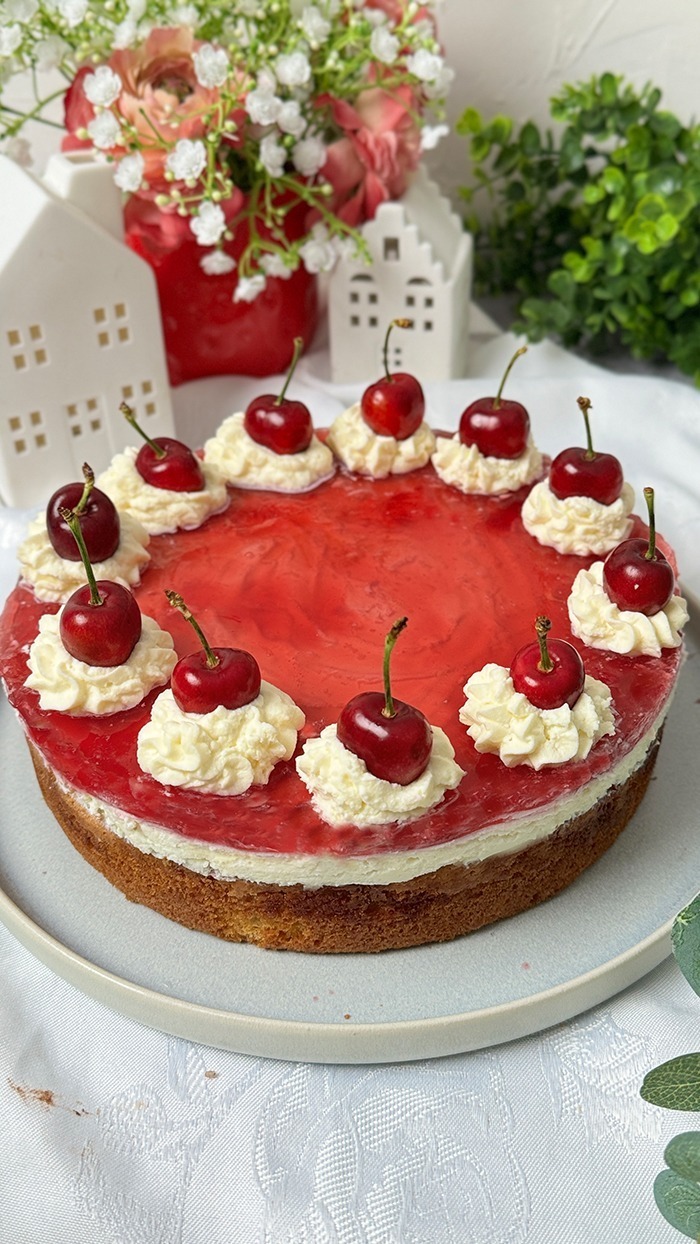 Auf dem Bild sieht man einen Rotkäppchen Kuchen, der auf einem Teller präsentiert wird. Der Kuchen hat eine helle, leicht goldene Biskuitbasis, die mit einer Schicht Sahne oder Pudding und einer roten Geleeschicht bedeckt ist. Am Rand des Kuchens befinden sich Sahnetupfen, auf denen jeweils eine frische, glänzende Kirsche mit Stiel platziert ist. Die Sahnetupfen sind gleichmäßig rund um den Kuchen verteilt. 