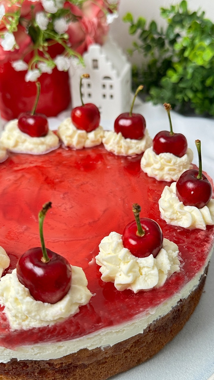 Auf dem Bild sieht man einen Rotkäppchen Kuchen, der auf einem Teller präsentiert wird. Der Kuchen hat eine helle, leicht goldene Biskuitbasis, die mit einer Schicht Sahne oder Pudding und einer roten Geleeschicht bedeckt ist. Am Rand des Kuchens befinden sich Sahnetupfen, auf denen jeweils eine frische, glänzende Kirsche mit Stiel platziert ist. Die Sahnetupfen sind gleichmäßig rund um den Kuchen verteilt. 