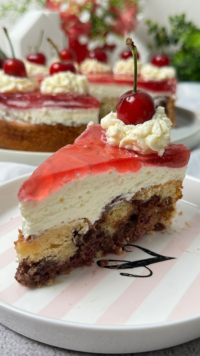 Auf dem Bild sieht man einen Rotkäppchen Kuchen, der auf einem Teller präsentiert wird. Der Kuchen hat eine helle, leicht goldene Biskuitbasis, die mit einer Schicht Sahne oder Pudding und einer roten Geleeschicht bedeckt ist. Am Rand des Kuchens befinden sich Sahnetupfen, auf denen jeweils eine frische, glänzende Kirsche mit Stiel platziert ist. Die Sahnetupfen sind gleichmäßig rund um den Kuchen verteilt. 