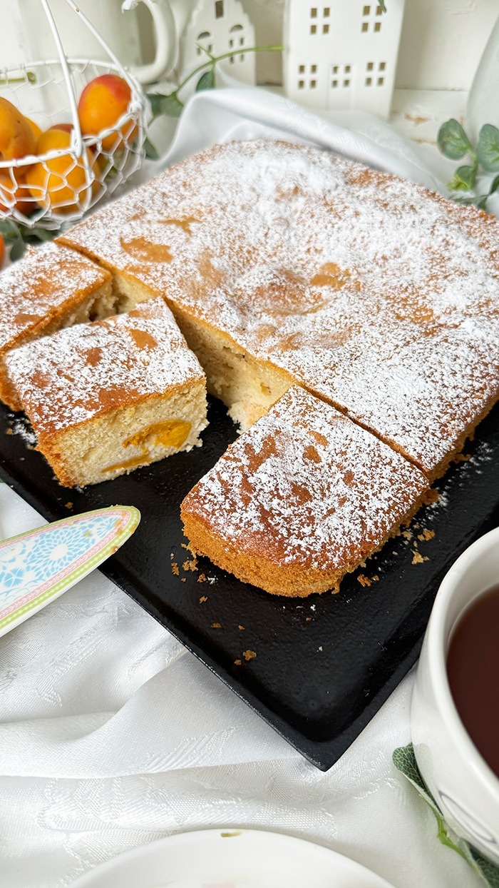 Ein köstlicher, rechteckiger Aprikosenkuchen, bestäubt mit Puderzucker, ist auf einem Teller und einer schwarzen Kuchenplatte angerichtet