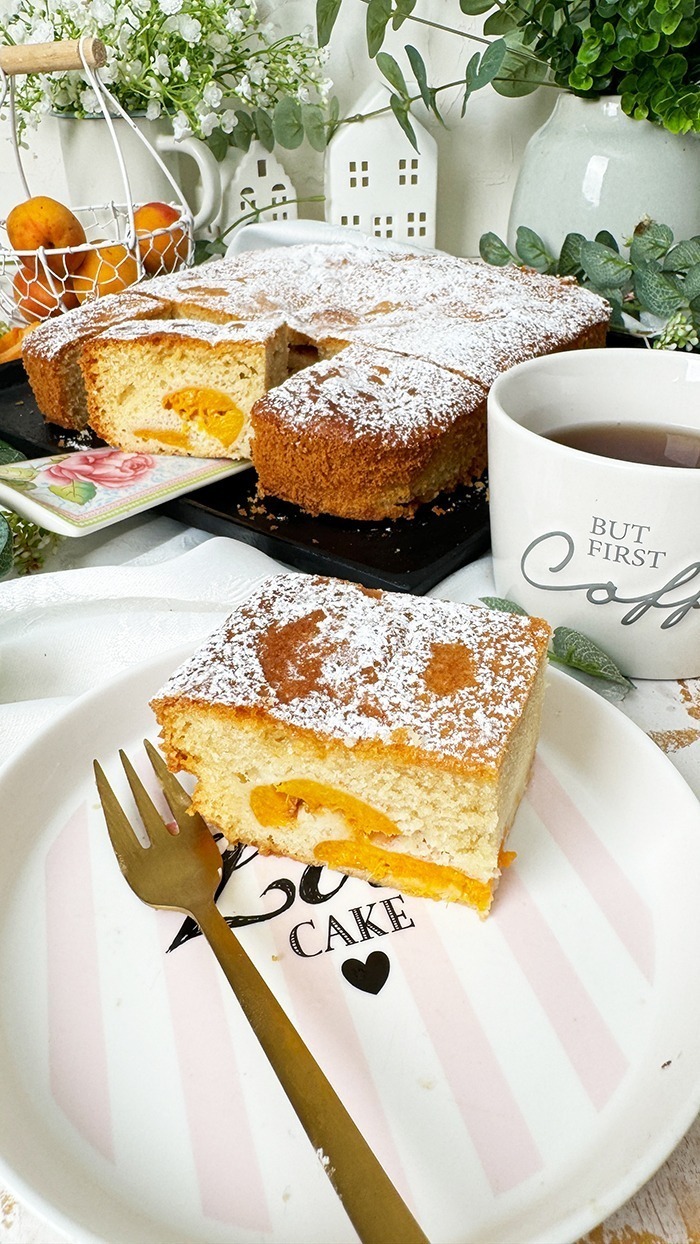 Ein köstlicher, rechteckiger Aprikosenkuchen, bestäubt mit Puderzucker, ist auf einem Teller und einer schwarzen Kuchenplatte angerichtet