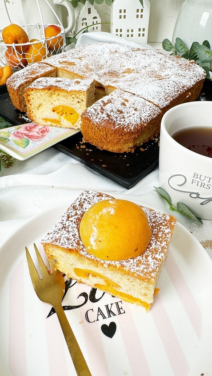 Ein köstlicher, rechteckiger Aprikosenkuchen, bestäubt mit Puderzucker, ist auf einem Teller und einer schwarzen Kuchenplatte angerichtet