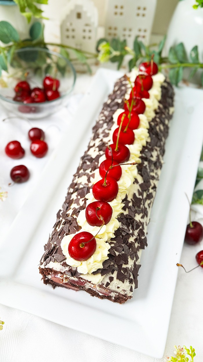 Das Bild zeigt eine Schwarzwälder Kirschrolle mit Biskuitteig, gefüllt mit Sahne und Kirschen, bedeckt mit Schokoladenraspeln und dekoriert mit Kirschen. Zwei Scheiben der Rolle liegen auf grauen Tellern mit goldenen Gabeln, umgeben von frischen Kirschen und grünen Blättern