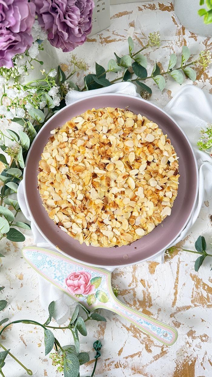 Auf dem Bild sieht man ein Stück Schwedische Mandeltorte, das auf einem grauen Teller angerichtet ist. Die Torte besteht aus mehreren Schichten: ein luftiger Mandelboden, dazwischen eine reichhaltige Vanillebuttercreme und obenauf eine Schicht gerösteter Mandelblättchen.