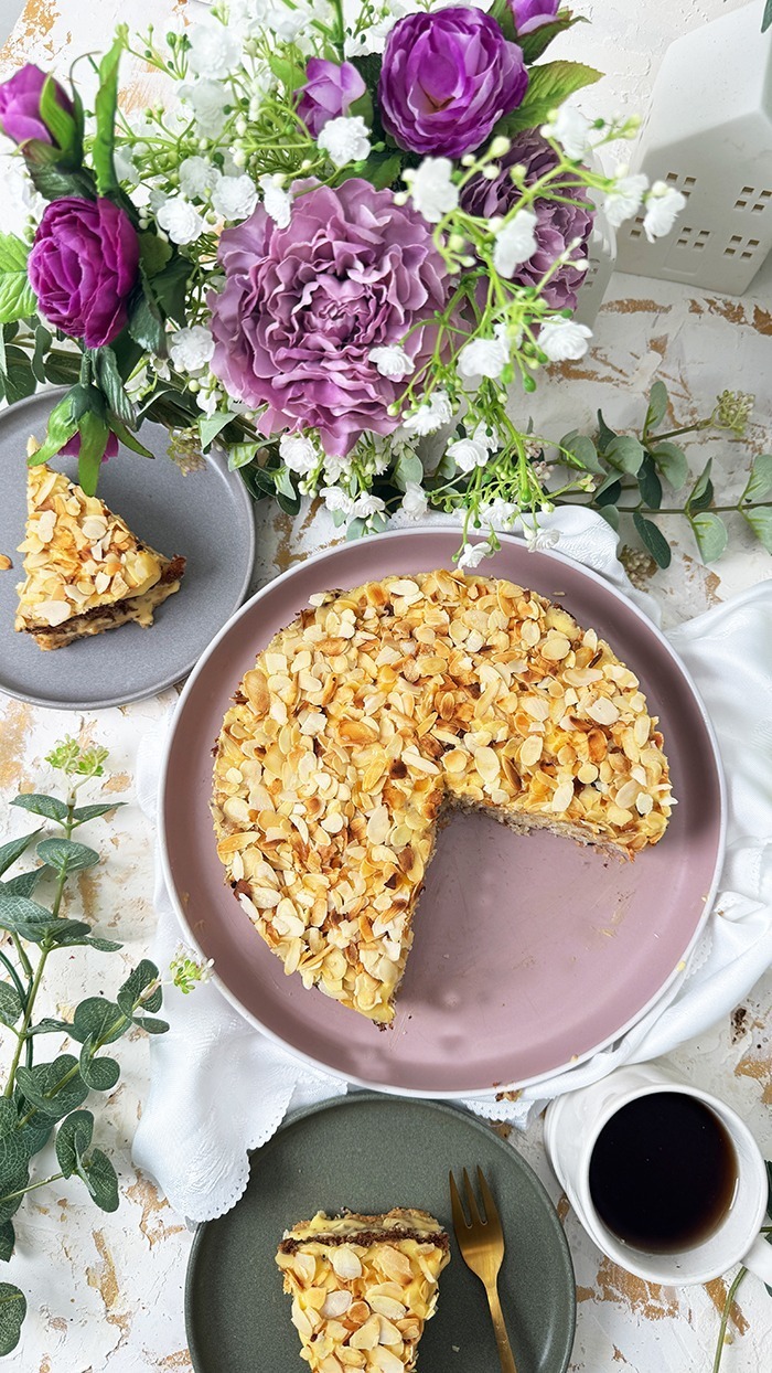 Auf dem Bild sieht man ein Stück Schwedische Mandeltorte, das auf einem grauen Teller angerichtet ist. Die Torte besteht aus mehreren Schichten: ein luftiger Mandelboden, dazwischen eine reichhaltige Vanillebuttercreme und obenauf eine Schicht gerösteter Mandelblättchen.