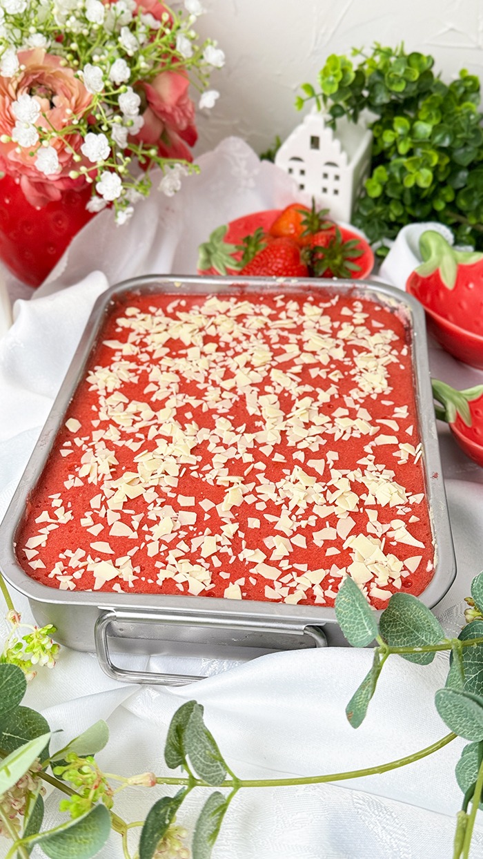 Dieses Bild zeigt ein Spaghettieis-Tiramisu. Das Dessert befindet sich in einer Backform, aus der ein quadratisches Stück herausgeschnitten und auf einem Teller angerichtet wurde, bereit zum Verzehr. Die oberste Schicht ist mit einer Erdbeerglasur und weißen Schokoladenraspeln garniert. Das Setting ist hell und mit Blumen und kleinen Pflanzen dekoriert, was auf ein Frühlings- oder Sommerthema hindeutet.