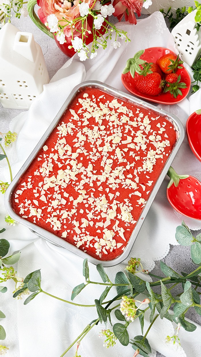 Dieses Bild zeigt ein Spaghettieis-Tiramisu. Das Dessert befindet sich in einer Backform, aus der ein quadratisches Stück herausgeschnitten und auf einem Teller angerichtet wurde, bereit zum Verzehr. Die oberste Schicht ist mit einer Erdbeerglasur und weißen Schokoladenraspeln garniert. Das Setting ist hell und mit Blumen und kleinen Pflanzen dekoriert, was auf ein Frühlings- oder Sommerthema hindeutet.