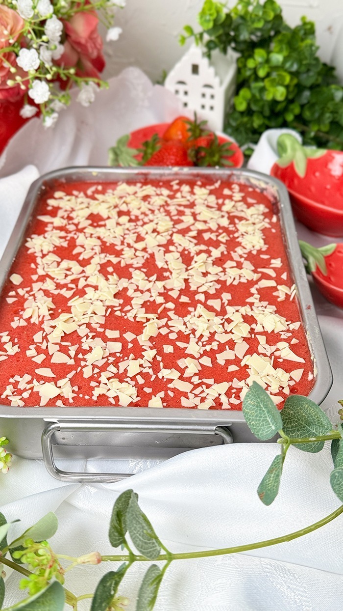 Dieses Bild zeigt ein Spaghettieis-Tiramisu. Das Dessert befindet sich in einer Backform, aus der ein quadratisches Stück herausgeschnitten und auf einem Teller angerichtet wurde, bereit zum Verzehr. Die oberste Schicht ist mit einer Erdbeerglasur und weißen Schokoladenraspeln garniert. Das Setting ist hell und mit Blumen und kleinen Pflanzen dekoriert, was auf ein Frühlings- oder Sommerthema hindeutet.