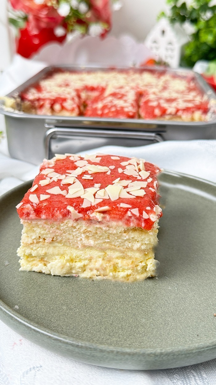 Dieses Bild zeigt ein Spaghettieis-Tiramisu. Das Dessert befindet sich in einer Backform, aus der ein quadratisches Stück herausgeschnitten und auf einem Teller angerichtet wurde, bereit zum Verzehr. Die oberste Schicht ist mit einer Erdbeerglasur und weißen Schokoladenraspeln garniert. Das Setting ist hell und mit Blumen und kleinen Pflanzen dekoriert, was auf ein Frühlings- oder Sommerthema hindeutet.