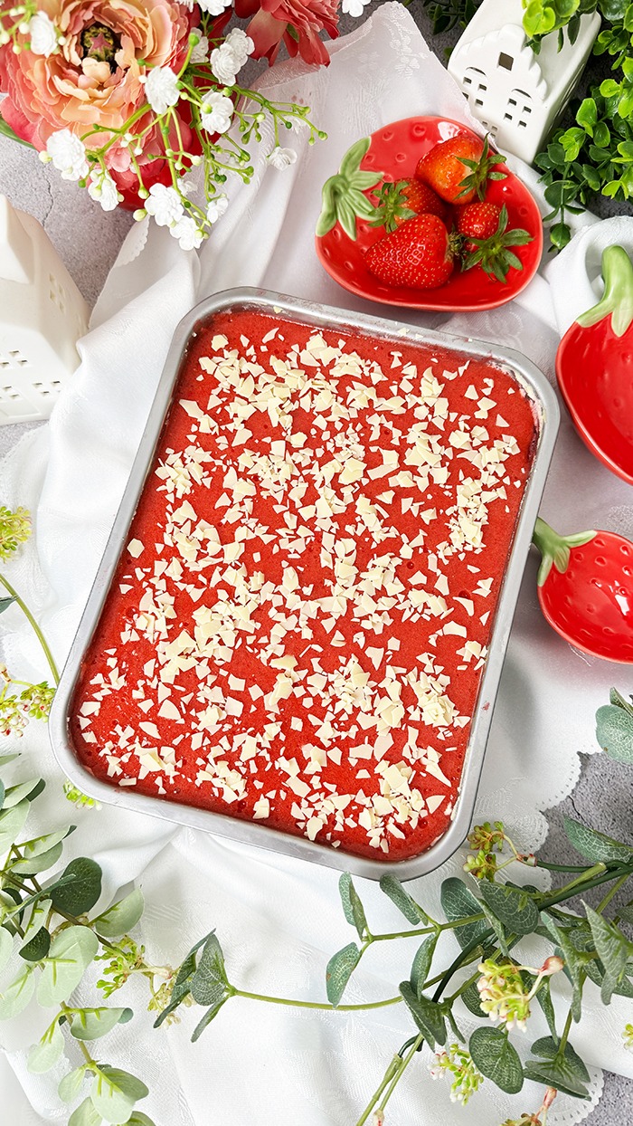 Dieses Bild zeigt ein Spaghettieis-Tiramisu. Das Dessert befindet sich in einer Backform, aus der ein quadratisches Stück herausgeschnitten und auf einem Teller angerichtet wurde, bereit zum Verzehr. Die oberste Schicht ist mit einer Erdbeerglasur und weißen Schokoladenraspeln garniert. Das Setting ist hell und mit Blumen und kleinen Pflanzen dekoriert, was auf ein Frühlings- oder Sommerthema hindeutet.