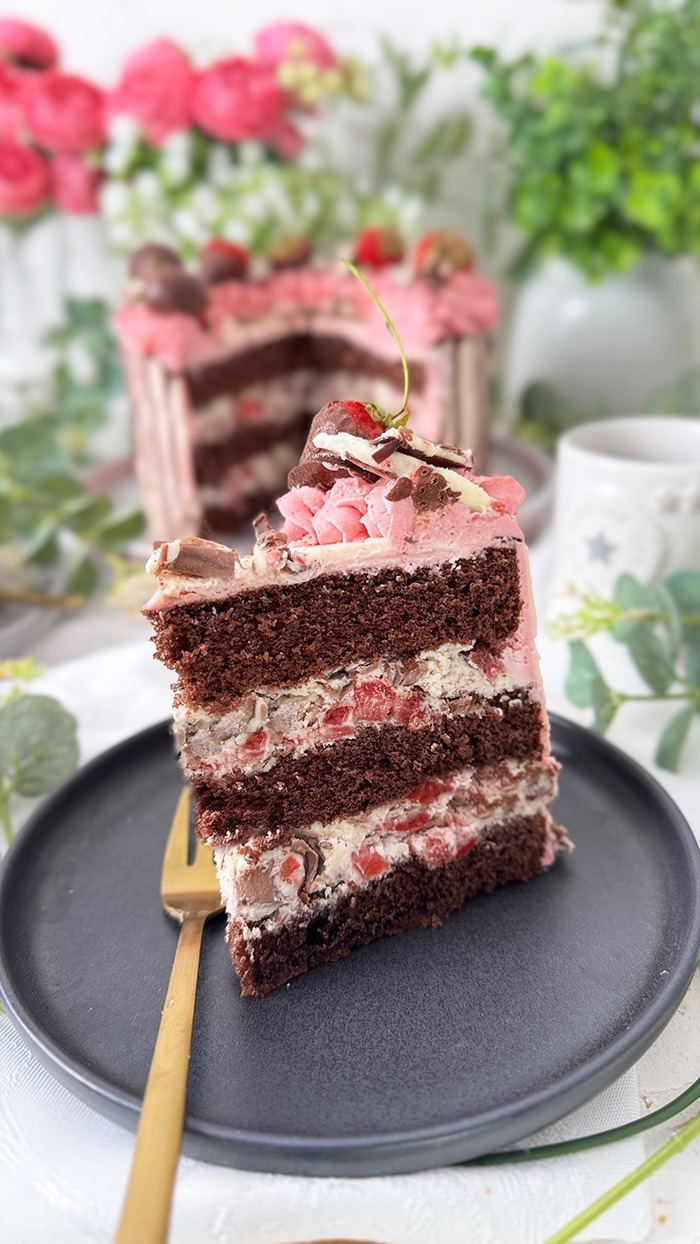 Das Bild zeigt eine zweischichtige Yogurette-Torte, dekoriert mit rosa Sahne, Schokoladen-Drip, in Schokolade getauchten Erdbeeren und Yogurette-Stückchen, umgeben von Blumen
