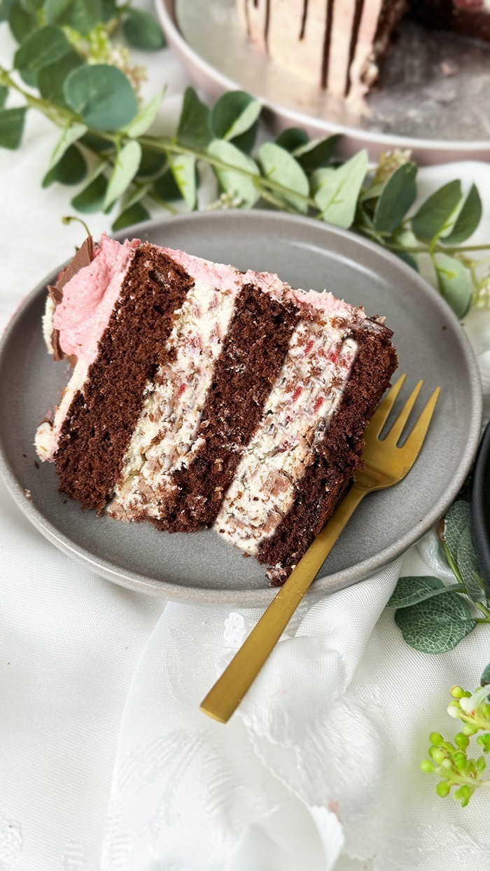 Das Bild zeigt eine zweischichtige Yogurette-Torte, dekoriert mit rosa Sahne, Schokoladen-Drip, in Schokolade getauchten Erdbeeren und Yogurette-Stückchen, umgeben von Blumen